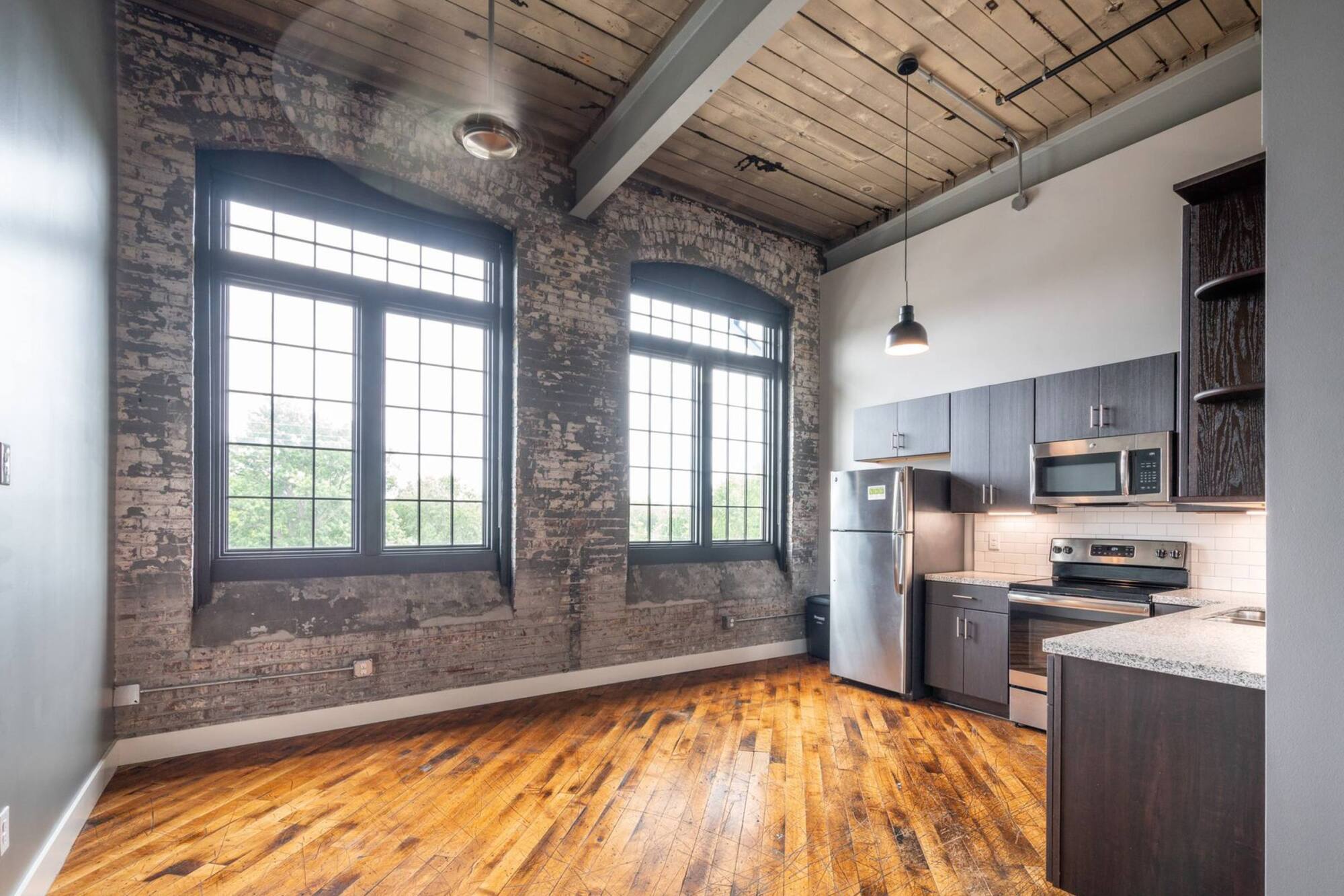 Modern loft kitchen with exposed brick, large industrial windows, wood ceiling, and hardwood floors.