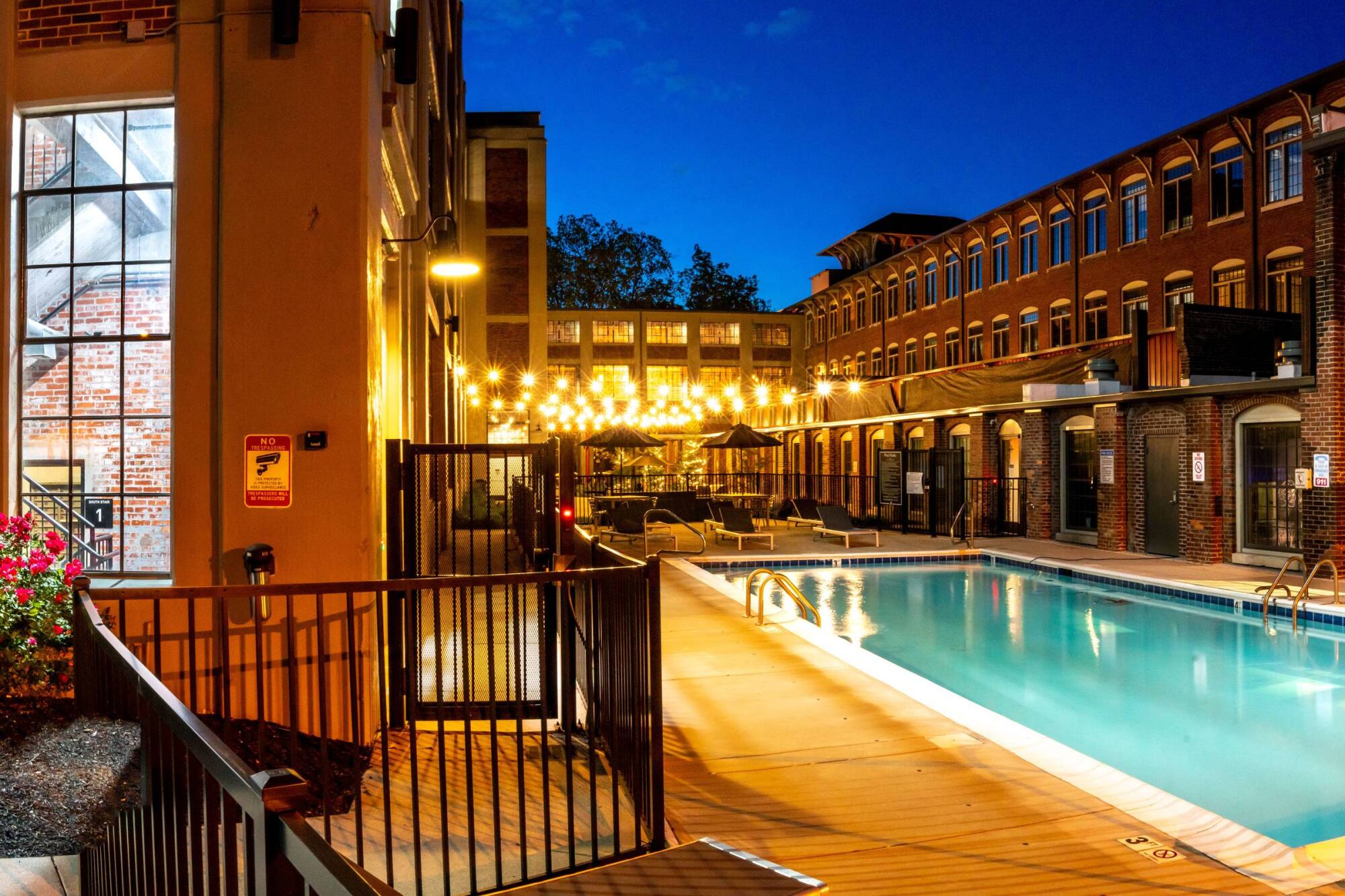 Outdoor pool at night, surrounded by brick buildings and string lights, with lounge chairs by the water.